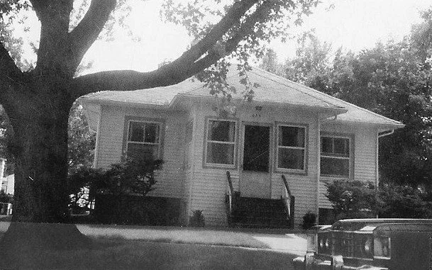 Black-and-white photo of a small single-story bungalow with a front porch, large tree in the yard, and a car parked at the curb.