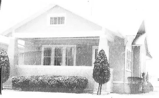 Black-and-white photo of a single-story bungalow with a covered front porch, low shrubs along the foundation, and a small conical tree by the porch.