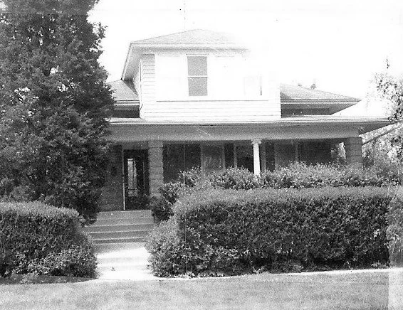 Black-and-white photo of a two-story house with a covered front porch, central steps, and tall hedges in front.