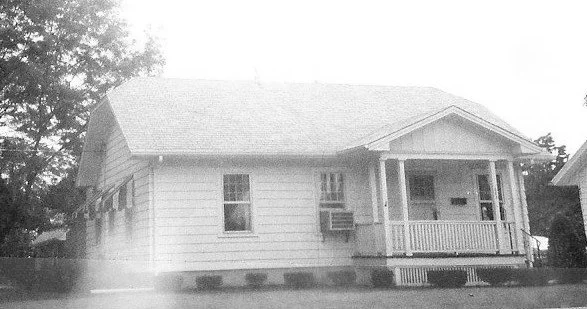 Small single-story wood-frame house with a covered front porch, steps, and a window air-conditioning unit; trees in the background.