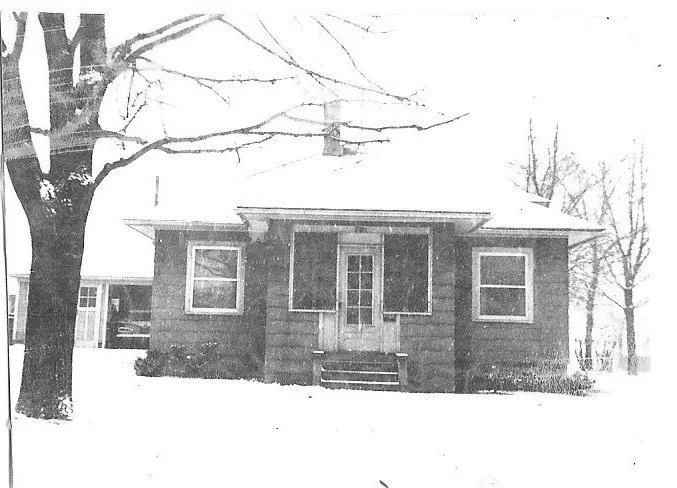 Black-and-white photo of a small single-story house with a centered front door and two windows, set in a snowy yard with a large bare tree to the left.