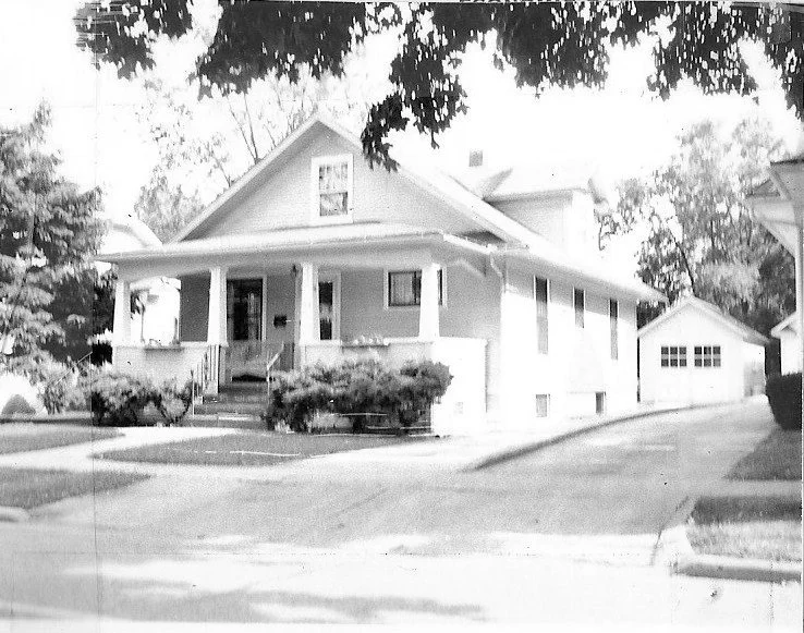 Black-and-white photo of a two-story house with a covered front porch, shrubs, a driveway leading to a detached garage, and tree branches overhead.