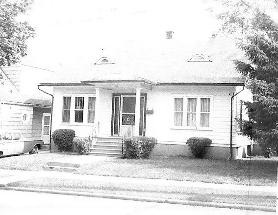 Black-and-white photo of a single-story house with a covered front porch, steps, shrubs, and a car partially visible at left.