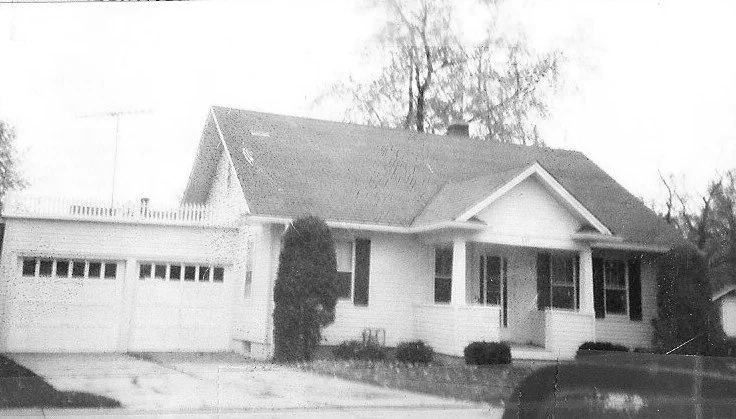 Black-and-white photo of a single-story suburban house with an attached two-car garage, front porch, shrubs, and trees in the background.
