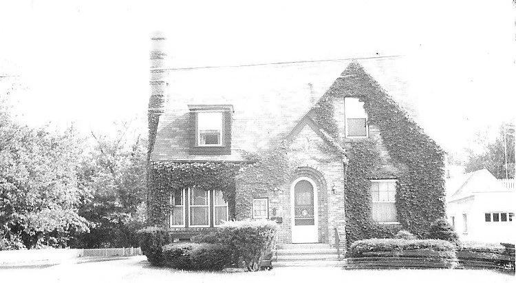 Ivy-covered brick house with arched front door, dormer windows, chimney and trimmed shrubs in front.