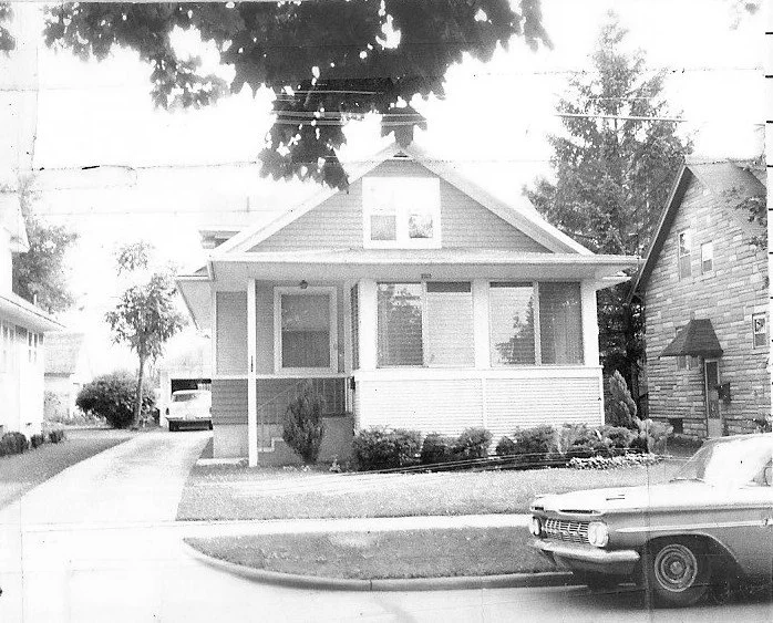 Black-and-white photo of a small single-story bungalow with an enclosed front porch, a vintage car parked on the street in front, and a driveway with a second car to the left.