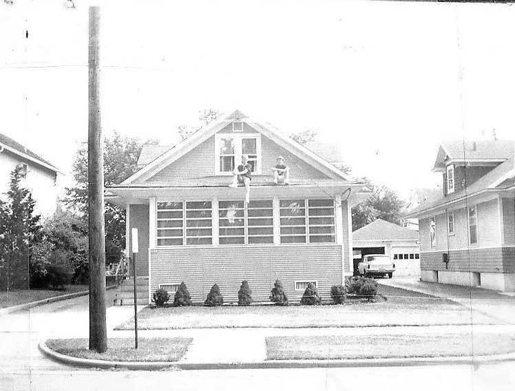 Black-and-white photo of a single-story bungalow with an enclosed front porch and two people sitting on the roof peak; driveway and parked car to the right.