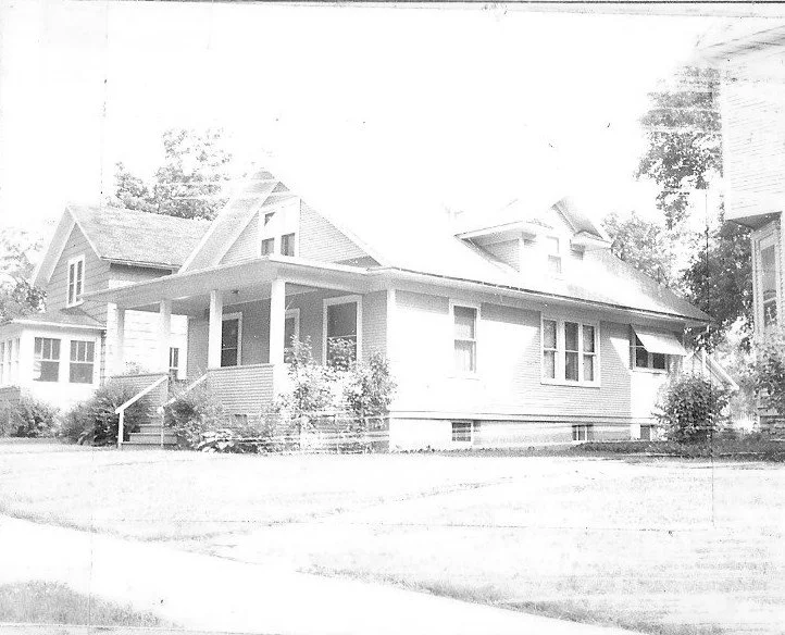 Black-and-white photo of a single-story bungalow with a covered front porch, lawn, shrubs, and neighboring houses visible.