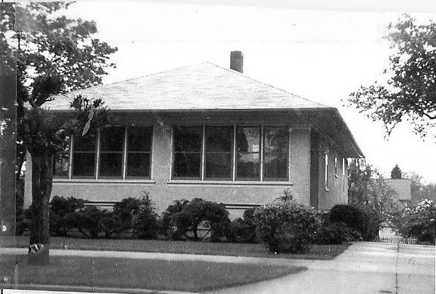 Black-and-white photo of a single-story bungalow with an enclosed front porch, low-pitched hip roof and chimney, surrounded by shrubs and trees with a sidewalk and driveway in front.
