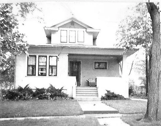 Black-and-white photo of a two-story Craftsman-style house with a front porch, central steps, shrubs, and trees on a residential street.