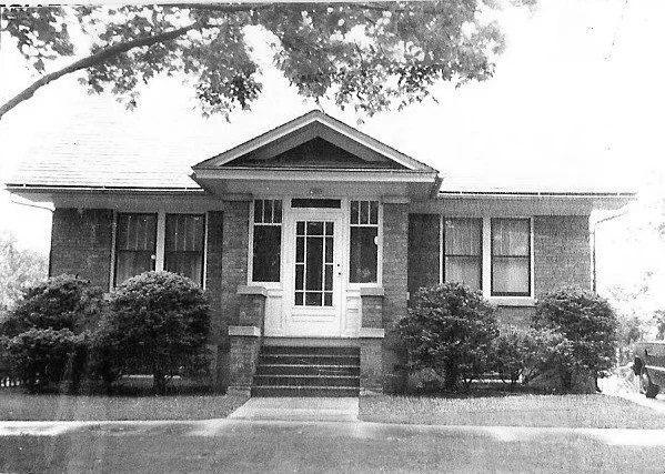 Black-and-white photo of a single-story brick bungalow with a centered enclosed front porch, steps up to a glass door, and shrubs lining the front.