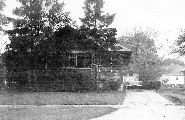 Black-and-white photo of a small bungalow partly hidden by tall trees, with a driveway leading to a vintage car parked at the side.