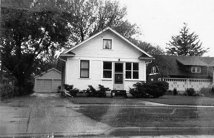 Black-and-white photo of a small single-story house with a screened front porch, detached garage to the left, driveway and trees in the background.