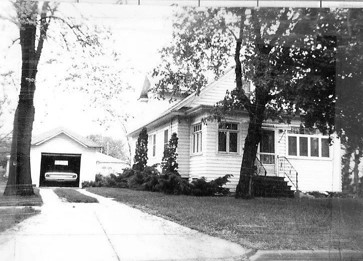 Black-and-white photo of a single-story wood-frame house with front steps and large trees, a driveway leading to a detached garage containing a vintage car.