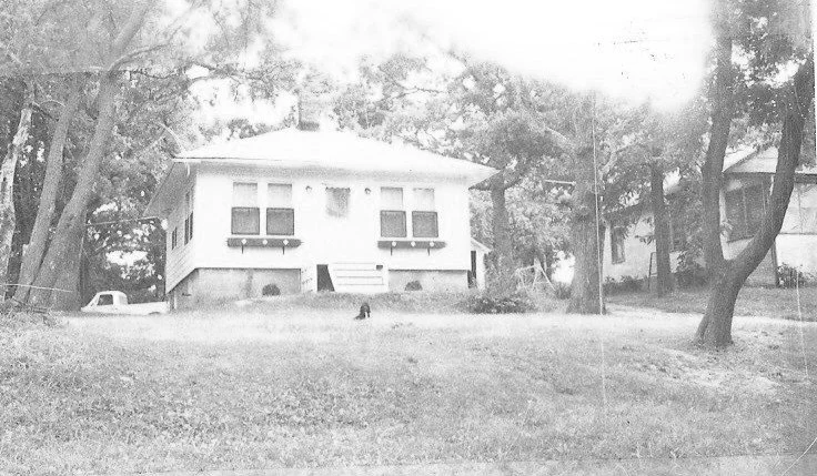 Small white bungalow on a grassy hill surrounded by large trees, with a parked pickup to the left and a small dark figure near the front steps.