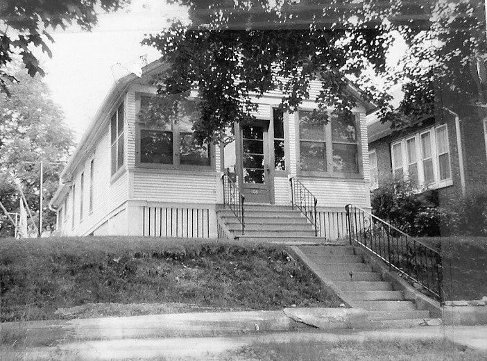 Black-and-white photo of a single-story bungalow with an enclosed front porch, central entrance reached by concrete steps and iron railings, framed by overhanging tree branches.