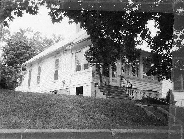Black-and-white photo of a single-story white clapboard house on a grassy slope with a screened front porch, metal-railed steps, and large tree branches overhead.