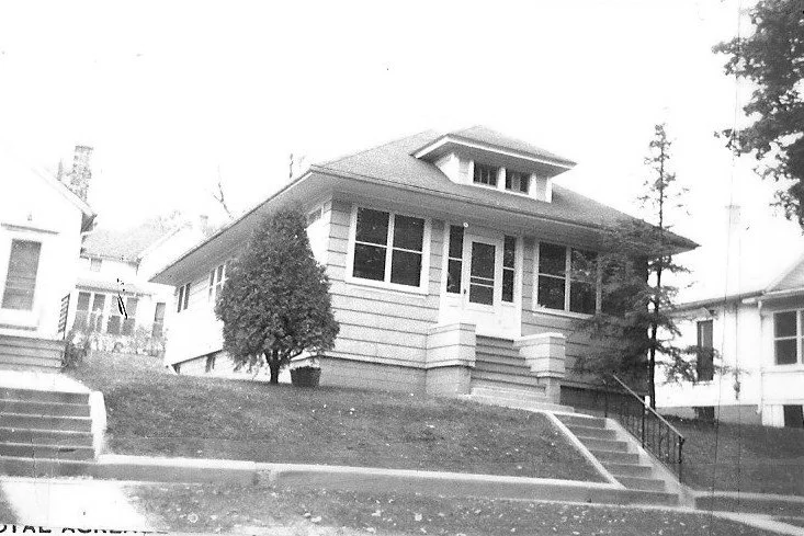 Black-and-white photo of a single-story bungalow with an enclosed front porch, centered front steps, a sloped lawn with small trees, and neighboring houses visible.