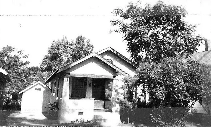 Black-and-white photo of a small bungalow with a covered front porch, detached garage to the left, and trees around the yard.