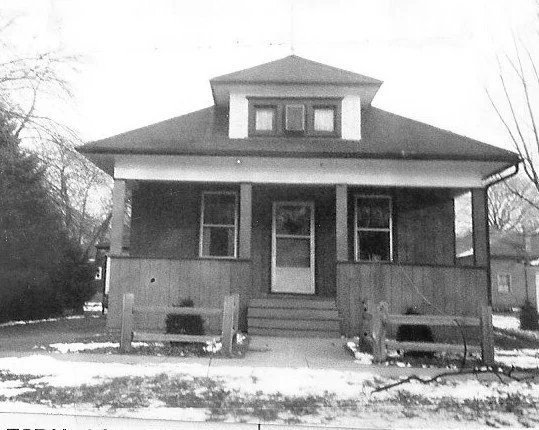 A front view of a small single-story bungalow with a covered porch, central door flanked by two windows, a dormer above, and patches of snow on the lawn.