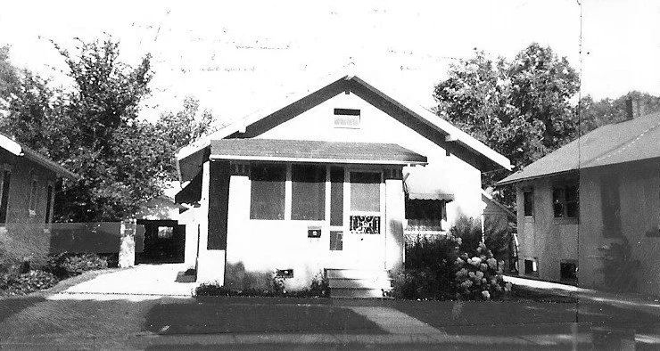 Black-and-white photo of a single-story bungalow with a small front porch, steps, neighboring houses, and trees in the background.