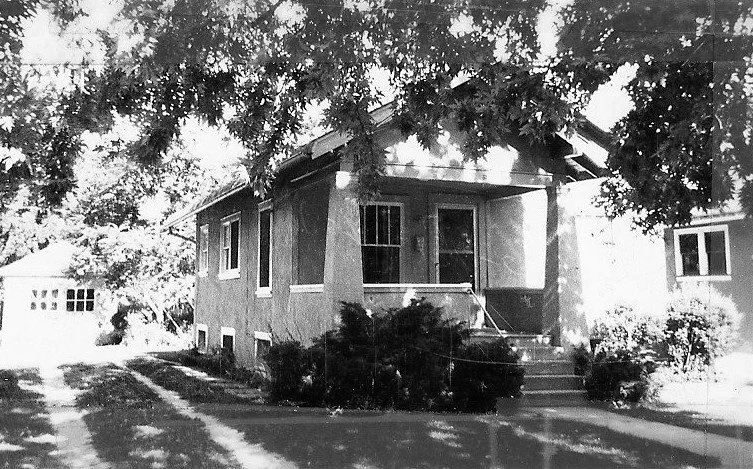 Black-and-white photo of a small bungalow with a front porch and steps, shrubs around the entrance, a large tree casting shadows overhead, and a detached garage visible to the left.