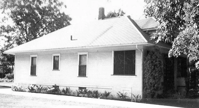 Black-and-white photo of a single-story white house with a low-pitched roof, several windows, shrubs along the foundation, and trees partially framing the right side.