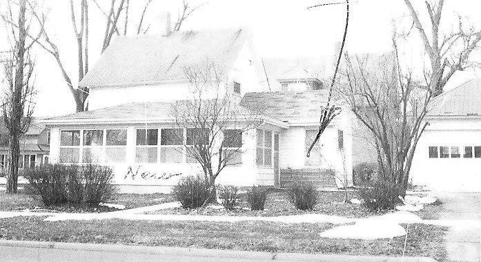 Black-and-white photo of a two-story house with an enclosed front porch, leafless trees, a driveway leading to a detached garage, and small patches of snow on the lawn.