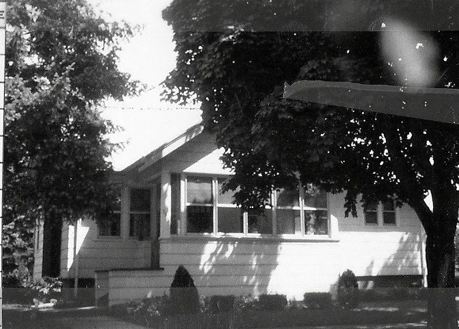 Black-and-white photo of a single-story white house with an enclosed front porch and large trees casting shadows in the yard.