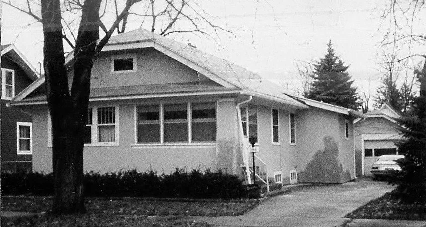 Black-and-white photo of a single-story bungalow with a large front tree, driveway leading to a detached garage and a parked car.