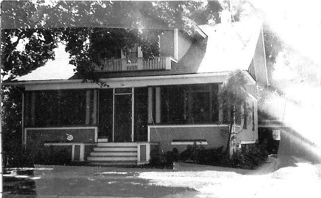 Black-and-white photo of a two-story house with a covered front porch, central steps, and a large tree casting shade.