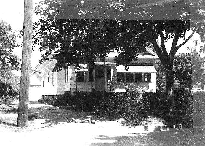 Black-and-white photo of a single-story house with a shaded front porch, large tree and a utility pole in the front yard.