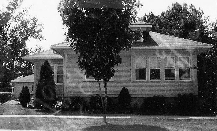 Black-and-white photo of a one-story house with a small tree and shrubs in front, viewed from the street.