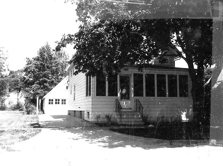 Black-and-white photo of a small house with an enclosed front porch, steps and railing, a large tree casting shade, and a driveway leading to a detached garage.