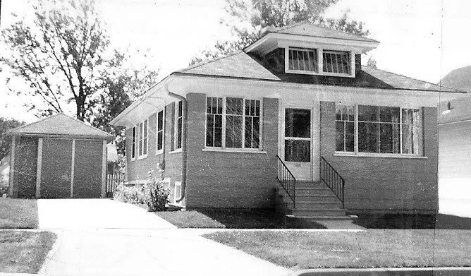 Front view of a brick bungalow with enclosed front porch, steps, driveway and detached garage.