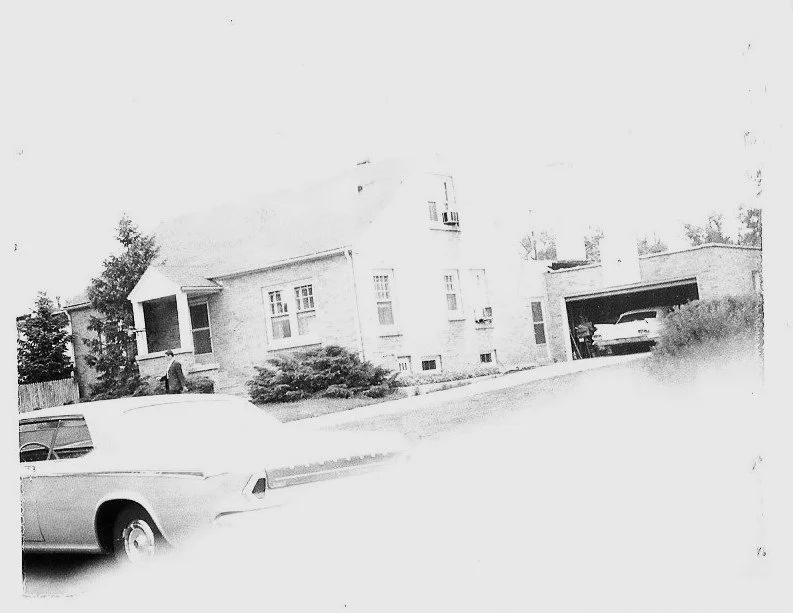 Faded black-and-white photo of a brick suburban house with a driveway and open garage containing a parked car; a second car is partially visible in the foreground.