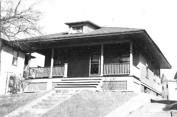 Black-and-white photo of a single-story bungalow with a wide covered front porch, steps up from the sidewalk, and a small dormer.