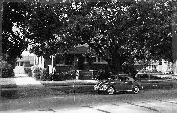Black-and-white photo of a tree-shaded bungalow with a person on the front walkway and a vintage Volkswagen Beetle parked on the street.