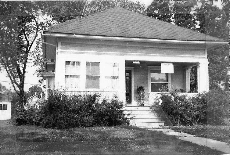 Black-and-white photo of a small bungalow with a covered front porch, concrete steps flanked by shrubs, lawn and trees, and a detached garage visible to the left.