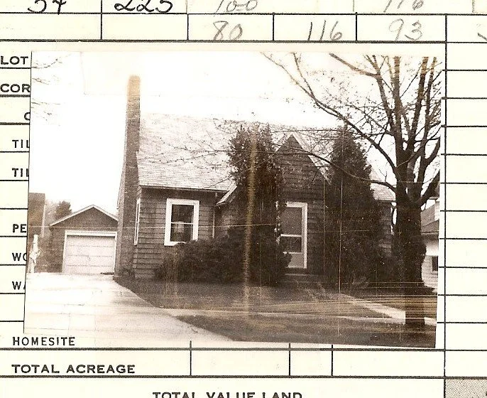 Black-and-white photo of a small gabled house with a tall chimney, detached garage, bare tree and shrubs, pasted onto a lined property form.