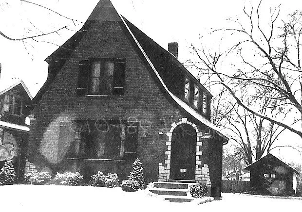 Snow-covered brick house with steep roof and arched stone entryway (house number 606), bare trees, and a small detached garage.