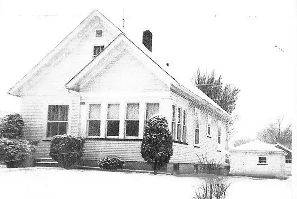 Front view of a white clapboard house with an enclosed porch, shrubs, and a detached garage in a snow-covered yard.
