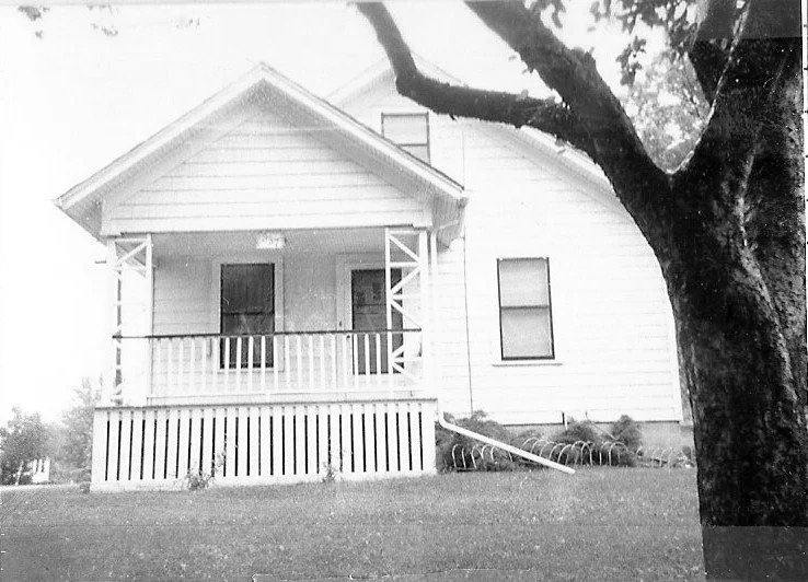 Black-and-white photo of a small wood-frame house with a covered front porch and railing, a large tree in the foreground at right.