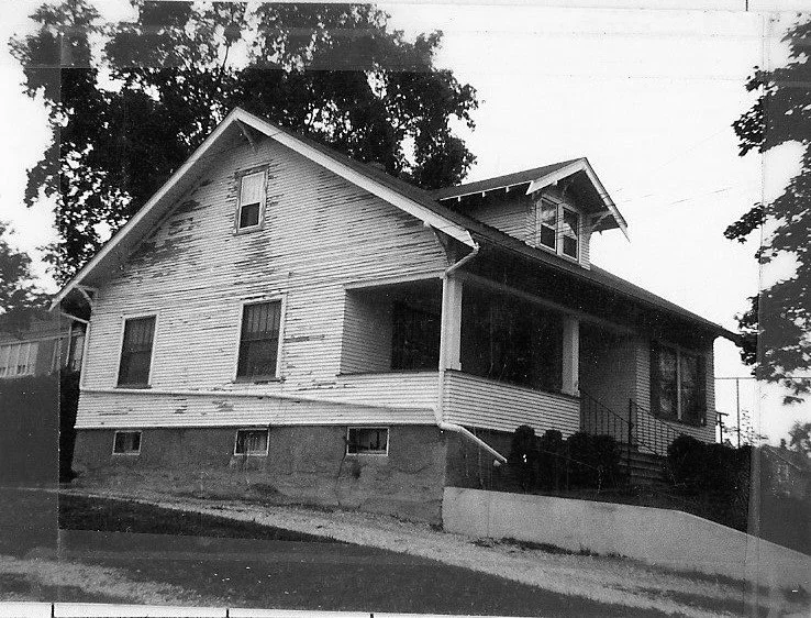 Black-and-white photo of an old wood-sided bungalow with a covered front porch and concrete foundation, set on a slightly sloped lot with trees.
