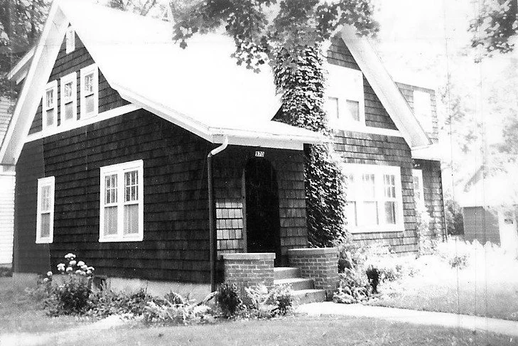 Black-and-white photo of a two-story shingled house with a small front porch, ivy on the entrance, and a lawn.