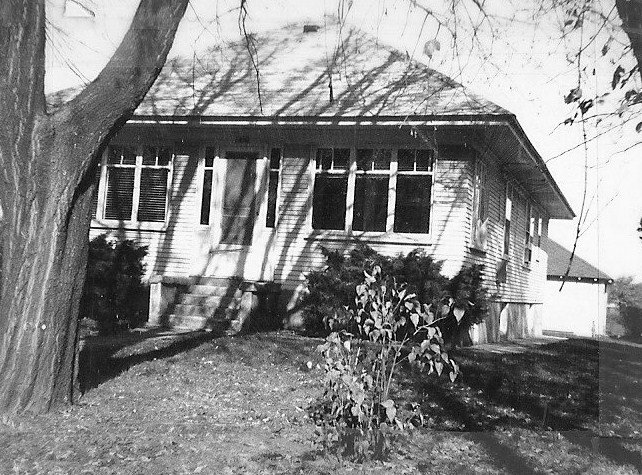 Black-and-white photo of a single-story bungalow with an enclosed front porch, large tree in the foreground, shrubs along the foundation, and a detached garage at the right.