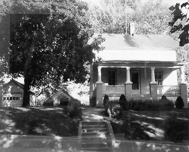 Black-and-white photo of a two-story house with a columned front porch, steps up a grassy yard, a large tree to the left, and a detached garage in the background.