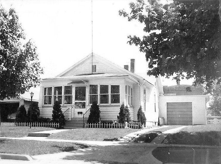 Black-and-white photo of a single-story house with an enclosed front porch, small picket fence, driveway to a detached garage, and trees framing the front yard.