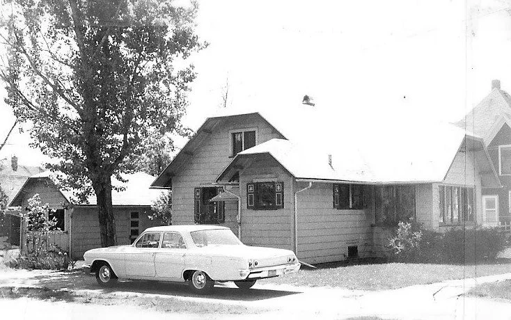 Black-and-white photo of a small bungalow with a large tree in the yard and a vintage 1960s sedan parked in front.