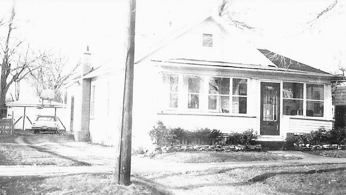 Small single-story house with an enclosed front porch, a driveway and parked car to the left, and a utility pole in the foreground.